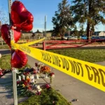 A police line with red balloons outside a school building. A couple of mourners are kneeling behind it.