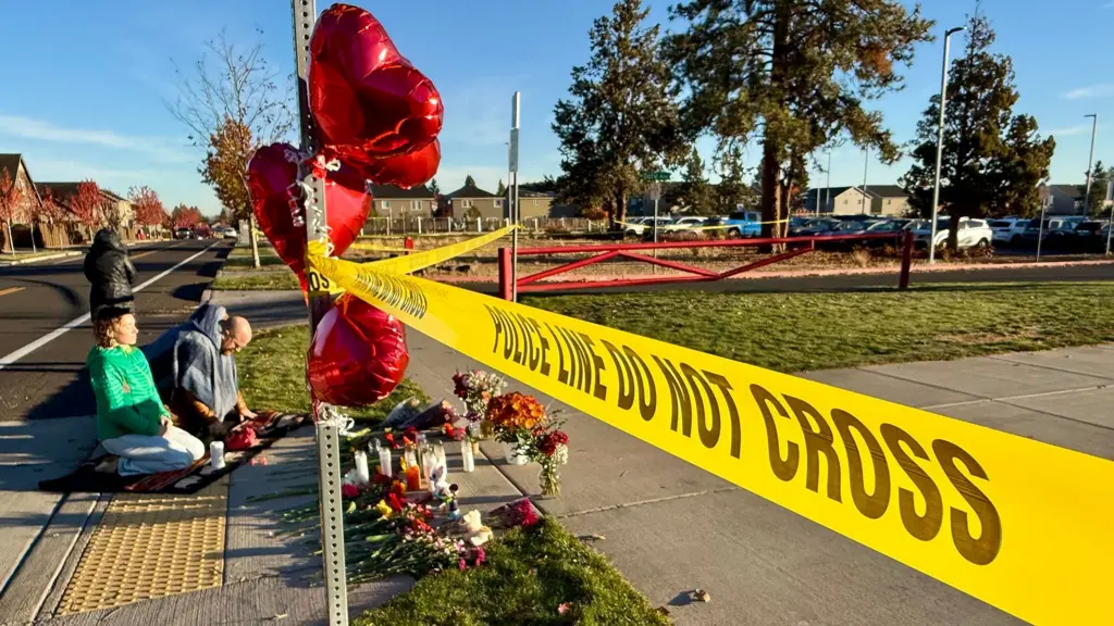 A police line with red balloons outside a school building. A couple of mourners are kneeling behind it.